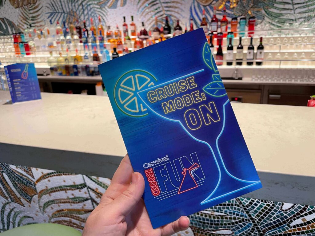 Hand holding a blue Carnival Cruise Line ‘Cruise Mode: ON’ menu in front of a brightly lit bar stocked with liquor bottles.”
