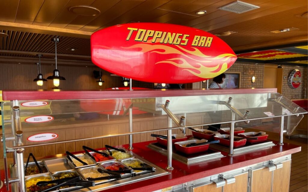 Bright red and yellow Toppings Bar sign over heated trays with various toppings and condiments.