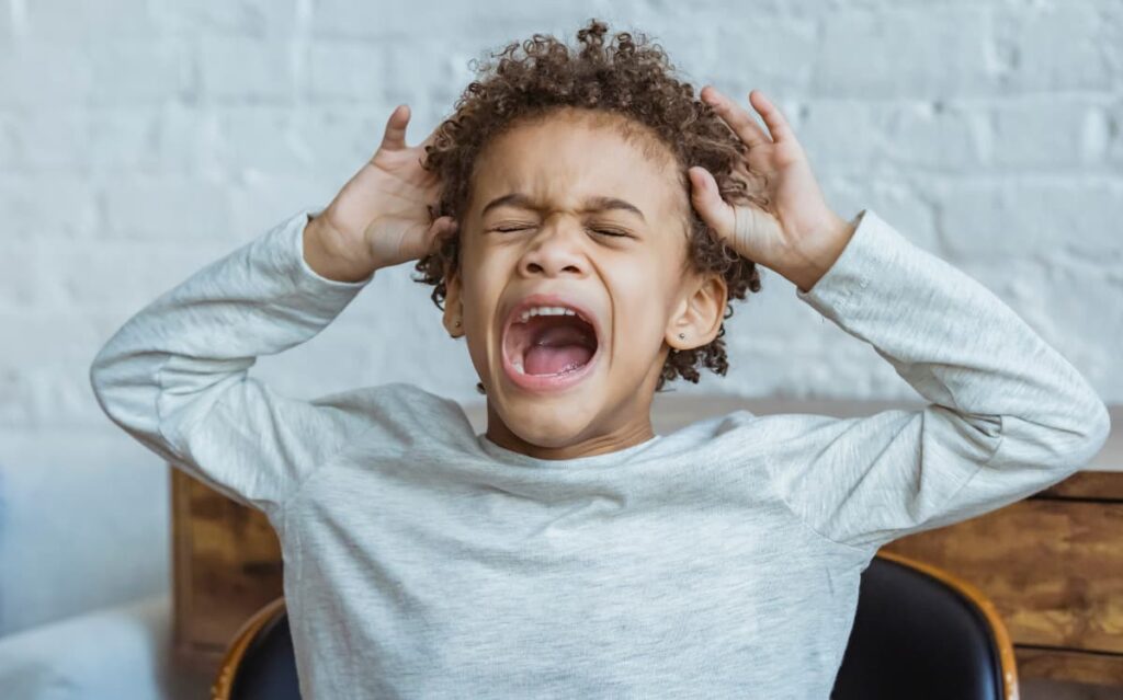 Young child with curly hair expressing frustration and yelling, hands on head.