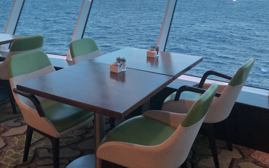 Dining table with green and tan chairs overlooking expansive blue ocean view through glass windows.