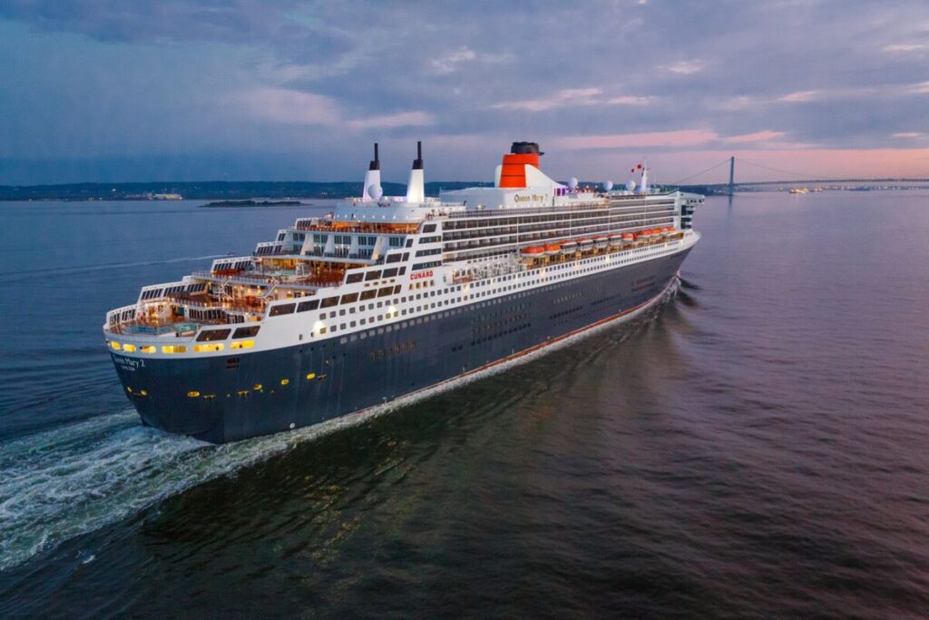 The majestic Cunard Queen Mary 2 cruise ship embarks on its voyage near New York, its distinctive red funnel and black hull illuminated during the early evening, with the Verrazzano-Narrows Bridge spanning the background.