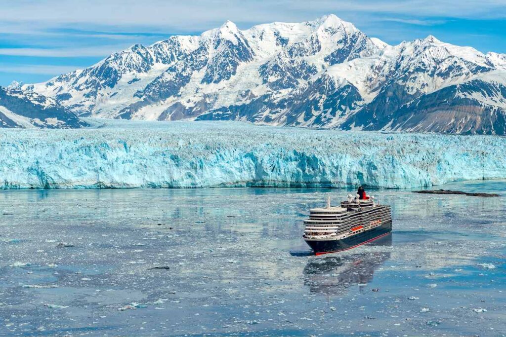 Cunard's Queen Elizabeth cruise ship is captured mid-journey amidst a serene sea of floating ice, with the majestic backdrop of a sprawling glacier and snow-capped mountains under a clear blue sky.
