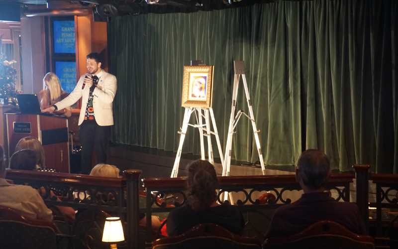 An art auction takes place on a cruise ship, featuring a well-dressed auctioneer presenting a framed piece of artwork displayed on an easel. The audience, seated comfortably, listens attentively as the auctioneer describes the artwork. The room is elegantly decorated with green curtains and soft lighting, creating a refined atmosphere for the event.