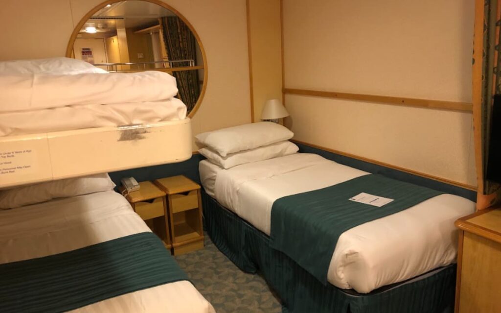 Cruise ship cabin interior with twin beds, nightstand, and round mirror, reflecting the compact and efficient design typical of cruise ship accommodations.