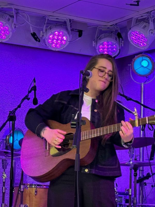 A solo performer playing an acoustic guitar in the 710 Club on P&O’s Iona, concentrating deeply on her music, illuminated by purple stage lights that create a relaxed and artistic atmosphere for the audience.