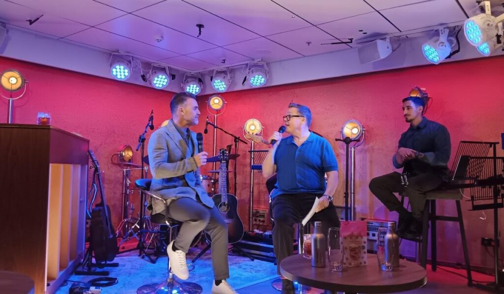 Three men engaged in a casual performance at the 710 Club on P&O’s Iona, with one speaking into a microphone and the others listening intently, in a cozy setting with vibrant stage lighting, musical instruments in the background, and a relaxed audience vibe.