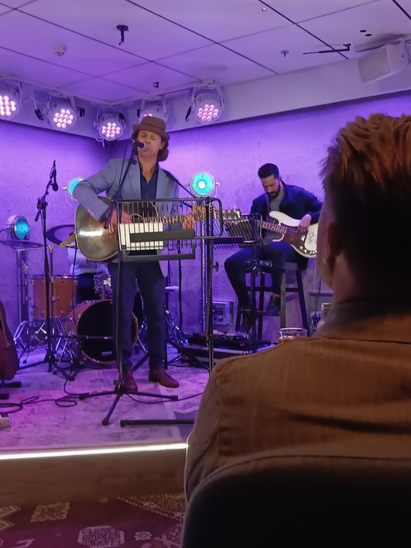 Live music session in the 710 Club on P&O’s Iona, with a musician in a hat playing a steel guitar and another on the bass guitar in the background, providing entertainment to the audience in an intimate club setting with warm lighting.
