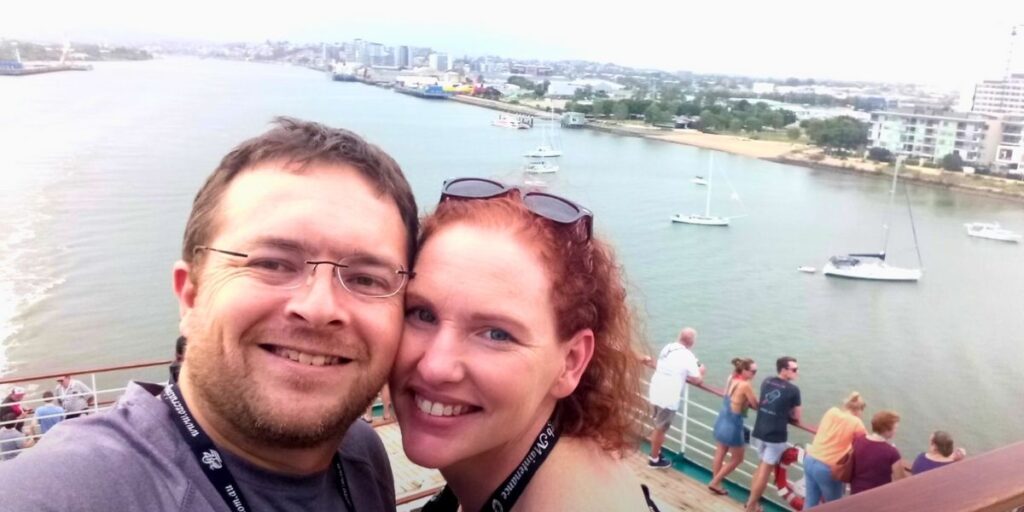 A smiling couple takes a selfie on the deck of a cruise ship as it departs a coastal city, with sailboats dotting the water and urban buildings in the background. Other passengers are gathered along the railing, enjoying the view of the shoreline and marina.