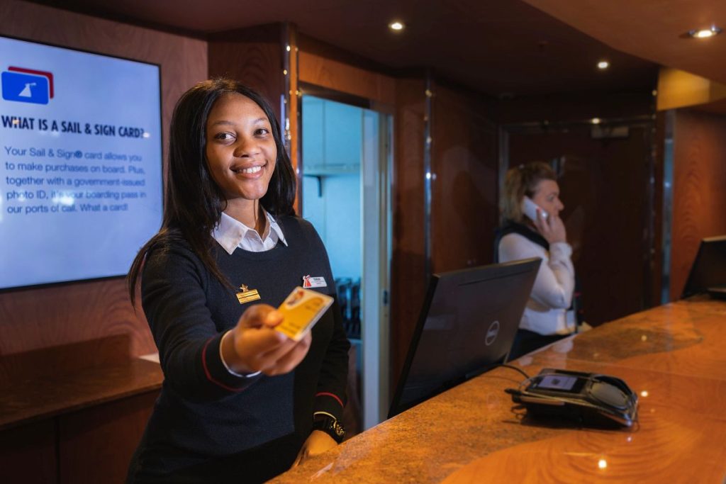 Friendly Carnival customer service representative extending a Sail & Sign card to guests, with an informative screen explaining the card's purpose in the background.