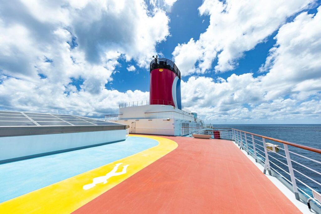 The image presents the jogging track on the Carnival Luminosa, marked by vibrant colors that stand out against the blue of the ocean and sky. The iconic red and blue funnel of the cruise ship looms in the background, and the track provides an inviting space for guests to exercise in the fresh sea air while enjoying the expansive view.