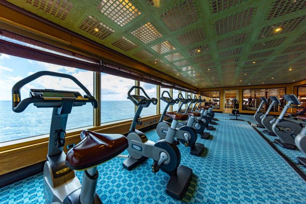 The image features the fitness center aboard the Carnival Luminosa, lined with a row of stationary bikes facing out toward the ocean. The spacious gym with its panoramic windows allows for an immersive workout experience with expansive sea views, under a decorative ceiling that adds a touch of elegance to the exercise environment.