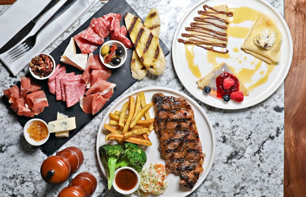 A beautifully arranged meal on a Carnival cruise ship features a variety of dishes. On one plate, there's a grilled steak served with French fries, broccoli, coleslaw, and a dipping sauce. Another plate showcases a selection of charcuterie, including slices of prosciutto, salami, cheese, olives, and toasted bread. A third plate offers a dessert of crepes drizzled with syrup and chocolate, accompanied by fresh berries. The spread is an enticing mix of savory and sweet, highlighting the gourmet dining experience available on the cruise.