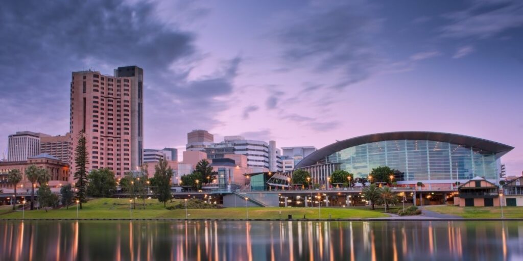 A view of the Adelaide city skyline at dusk featuring modern high-rise buildings and the glass-fronted Adelaide Convention Centre reflected in the calm waters of the River Torrens.