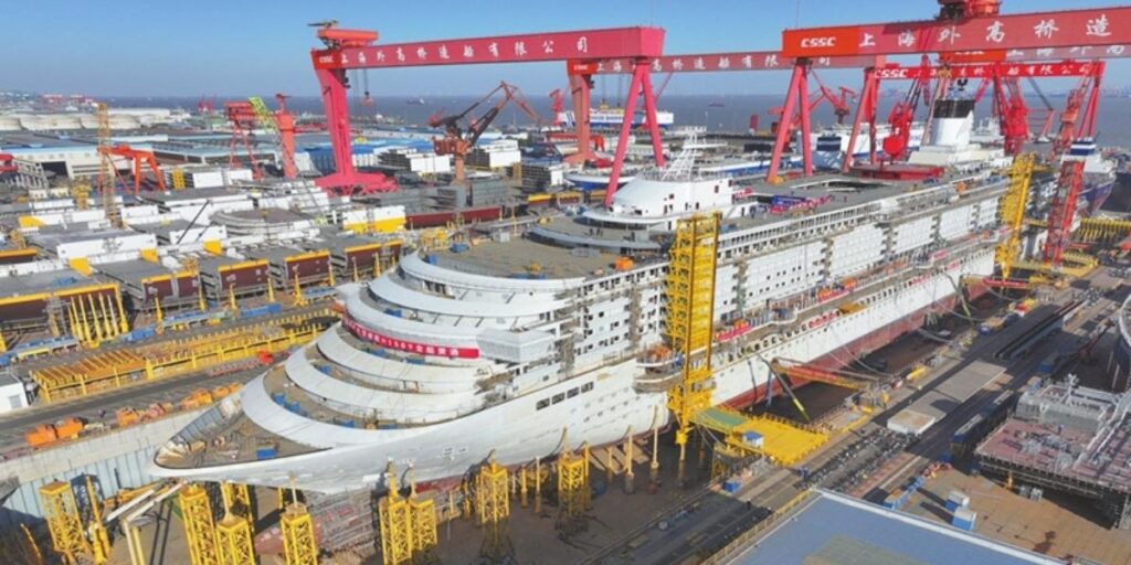 Aerial view of Adora Flora City under construction at a Chinese shipyard, showing the large cruise ship in dry dock surrounded by cranes, scaffolding, and industrial infrastructure.