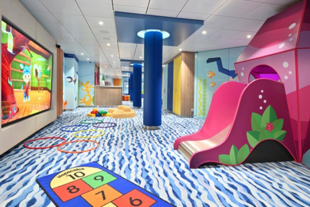 This image showcases the Adventure Ocean kids' play area on Icon of the Seas. The vibrant space is designed for young children, featuring colorful play structures, a slide, hopscotch mats, and interactive learning tools. The room is bright and playful, with animated characters displayed on a large screen and playful designs adorning the walls. The blue and white wavy carpet, along with the cheerful atmosphere, creates an engaging and safe environment for children to explore and enjoy activities.