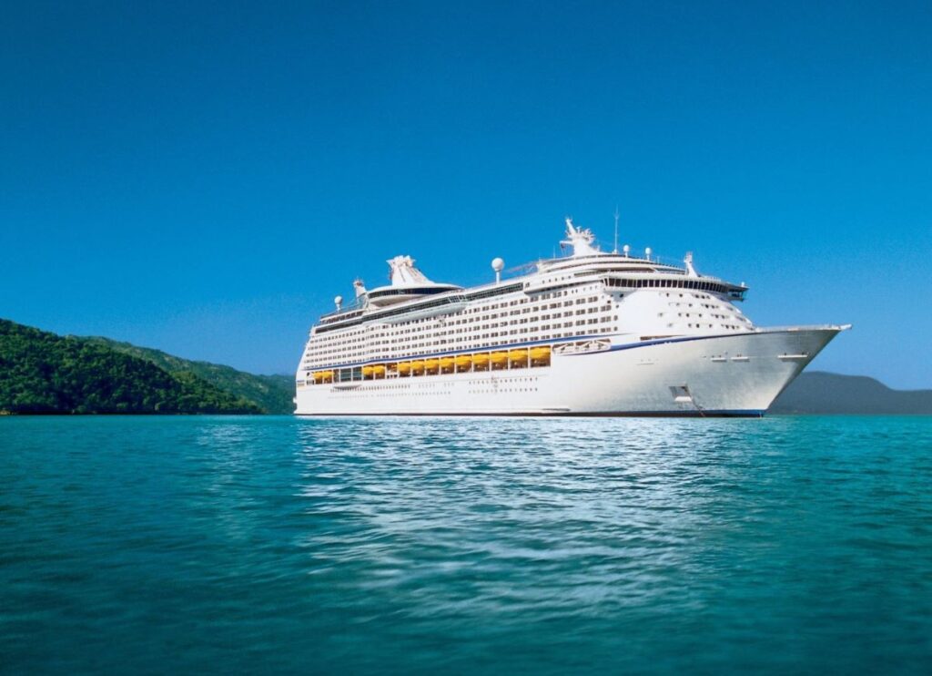 The Adventure of the Seas, a Royal Caribbean cruise ship, rests in a calm bay with crystal clear turquoise waters. Lush green hills frame the background, creating a tranquil and picturesque setting for this grand vessel under the bright, clear sky.