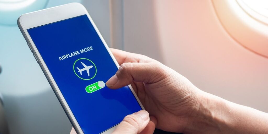 Close-up of a person’s hand switching a smartphone to airplane mode, with a blue screen displaying a plane icon and the word “ON” highlighted. The scene is set next to an airplane window, indicating the user is preparing for a flight.