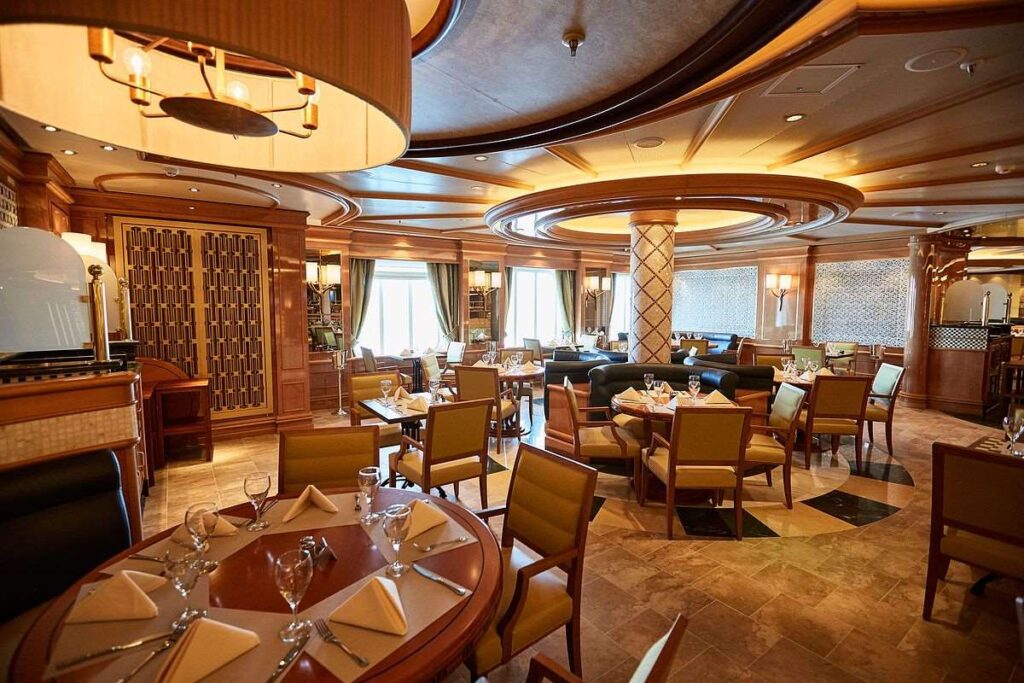 This image showcases Alfredo's Pizzeria aboard the Majestic Princess cruise ship, featuring an elegant and warmly lit dining space. The restaurant is adorned with polished wood finishes, cushioned chairs, and neatly set tables with folded napkins and glassware. The cozy ambiance, combined with refined decor and ample natural light streaming through large windows, creates an inviting atmosphere for guests to enjoy handcrafted pizzas and other Italian specialties.