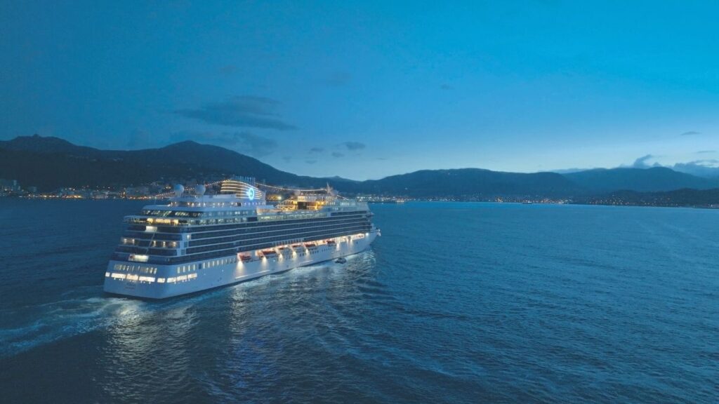 The Allura cruise ship from Oceania Cruises glides through calm waters at twilight, its elegant white exterior illuminated by warm onboard lights. The ship leaves a gentle wake behind as it sails past a coastal city, where twinkling lights dot the shoreline against a backdrop of rolling mountains. The serene blue sky and reflections on the water enhance the peaceful and luxurious ambiance of the voyage.