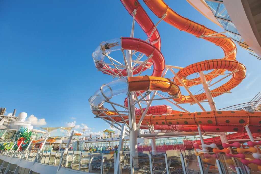 This image highlights the vibrant water slides aboard the Allure of the Seas cruise ship, featuring bold red and orange twists and turns against a bright blue sky. The slides are part of an expansive water park area designed for fun and excitement, with clear tubing sections offering a thrilling view of the surroundings. The dynamic design and sunny atmosphere showcase the family-friendly adventure available onboard.