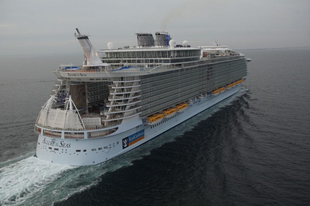 This image shows the Allure of the Seas, a massive Royal Caribbean cruise ship, sailing through calm open waters. The ship's innovative design features distinct neighborhoods, an open-air boardwalk, and multiple decks of staterooms and entertainment areas. Its impressive scale and modern amenities make it a standout vessel in the cruise industry.