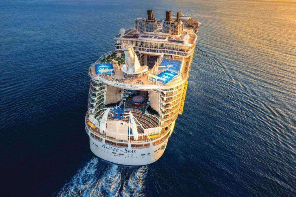 Aerial view of the Royal Caribbean Allure of the Seas, with its distinctive open-air aqua theater and multiple sports courts, gliding through the serene blue ocean during a warm, golden hour.