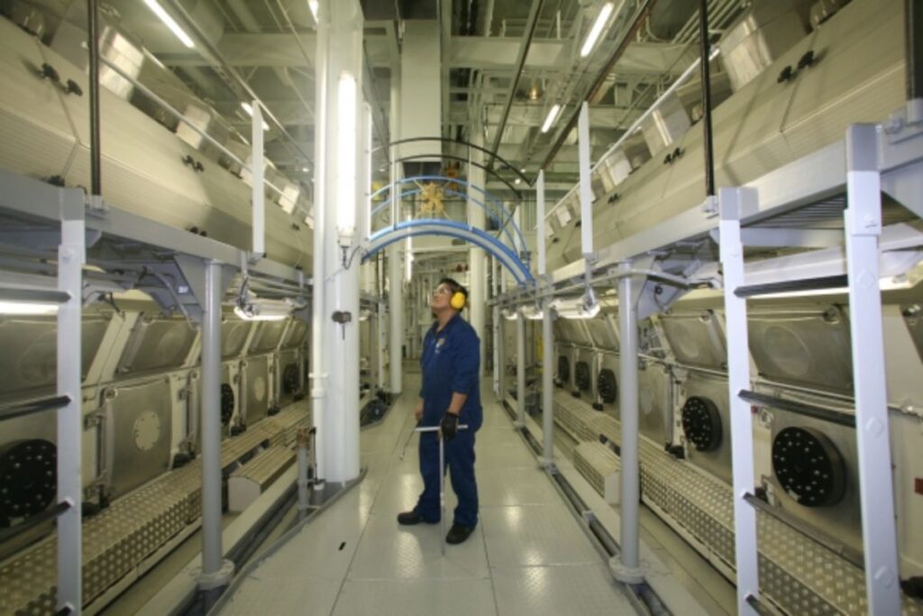 This image shows a person in protective gear, including earmuffs, standing in the engine room of the Allure of the Seas cruise ship. The room is lined with large, metallic machinery and intricate piping on both sides, giving the space an industrial and high-tech feel. Overhead, there is a metal walkway and bright lighting, highlighting the complexity of the equipment. The person appears to be inspecting or monitoring the machinery, indicating a focus on maintenance or operation.