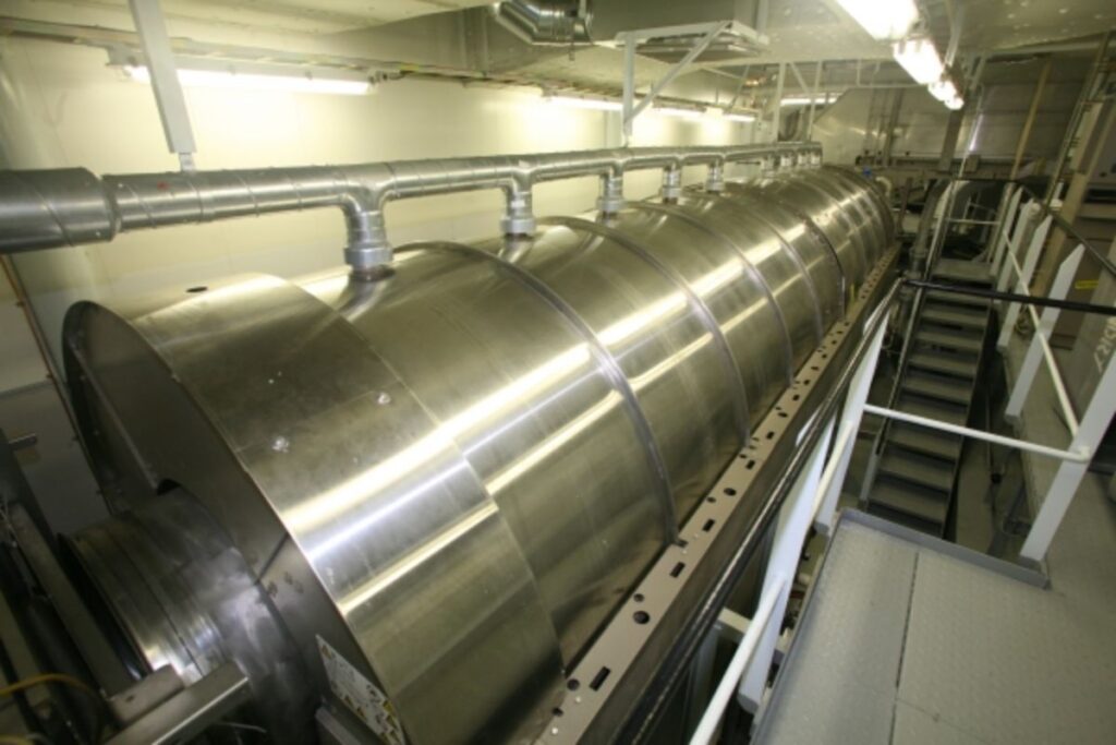 This image shows a large industrial machine in the laundry area of the Allure of the Seas cruise ship. The machinery appears to be a massive cylindrical washer or dryer, constructed from stainless steel and connected to overhead pipes, which likely help with ventilation or water supply. The setting is clean and well-lit, with metal stairs and walkways visible in the background, highlighting the ship's robust infrastructure for handling large-scale laundry operations.