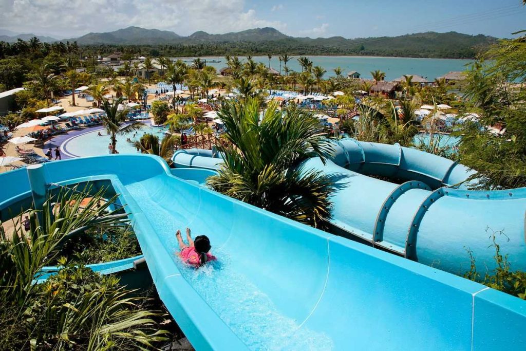 Child enjoying a water slide at Amber Cove, with a panoramic view of the resort's pools, palm trees, and the mountainous landscape of the Dominican Republic in the background.