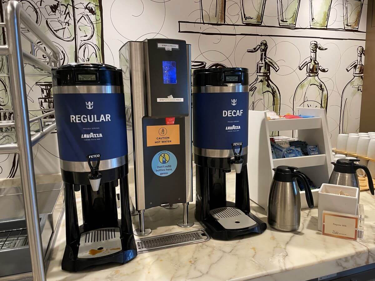 A Royal Caribbean beverage station featuring 'Regular' and 'Decaf' Lavazza coffee dispensers, a hot water station with a caution sign, and accompaniments including milk and sweeteners on a marble countertop.