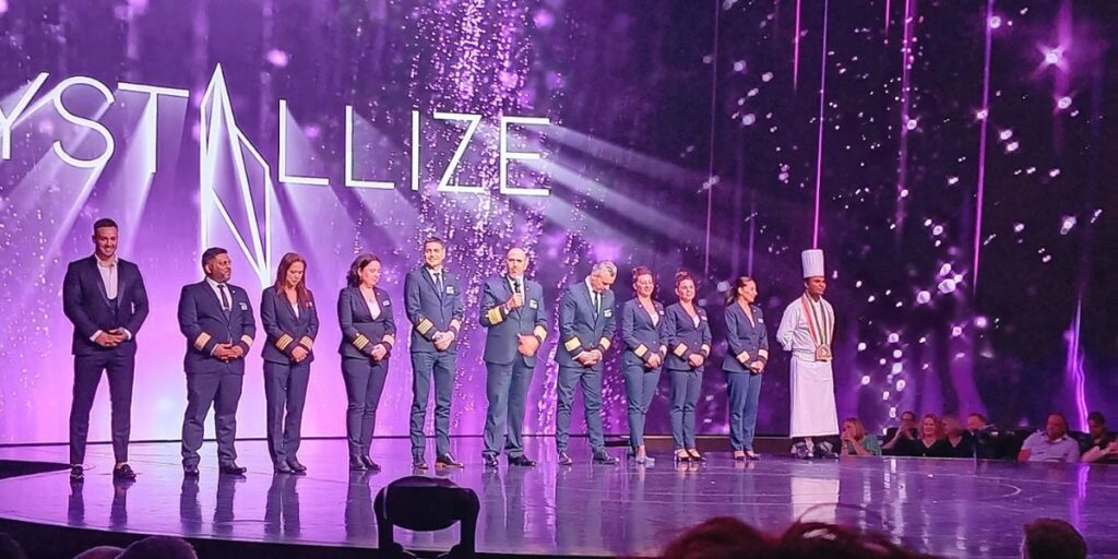 A lineup of crew members on the Celebrity Apex cruise ship, dressed in formal uniforms, standing on stage during an event called "Crystallize." The backdrop is a vibrant purple with sparkling effects, creating a celebratory atmosphere. One crew member is speaking into a microphone, while others, including a chef in a white uniform, stand attentively.