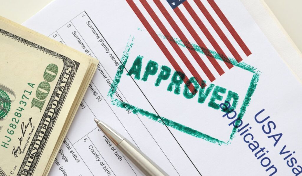 A close-up of a U.S. visa application with an 'APPROVED' stamp over the American flag, alongside a pen and a stack of one hundred dollar bills, indicating the successful completion of the visa application process.