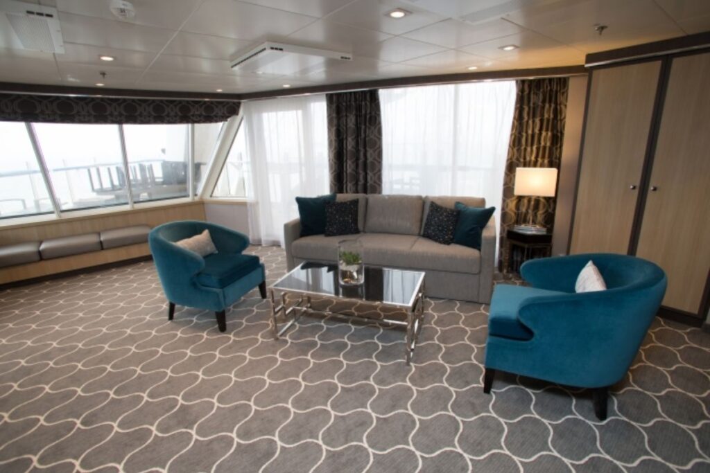 This image features the AquaTheater Suite on Harmony of the Seas, showcasing a spacious living area with stylish decor. The room includes a comfortable gray sofa, blue accent chairs, and a glass coffee table, all arranged on a patterned carpet. Large windows allow plenty of natural light, offering panoramic views. The suite exudes elegance and comfort, perfect for relaxing while enjoying the scenic ocean views.