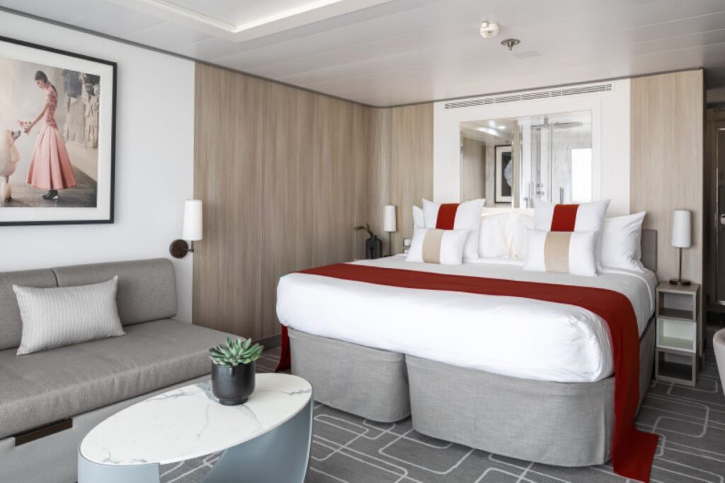 This image showcases an AquaClass Sky Suite aboard a Celebrity cruise ship, designed with a blend of modern elegance and comfort. The room features a spacious king-sized bed with crisp white linens and vibrant red accents, a cozy seating area, and a chic marble-topped coffee table. The neutral wood tones, soft lighting, and tasteful artwork create a sophisticated and tranquil atmosphere for a luxurious cruise experience.
