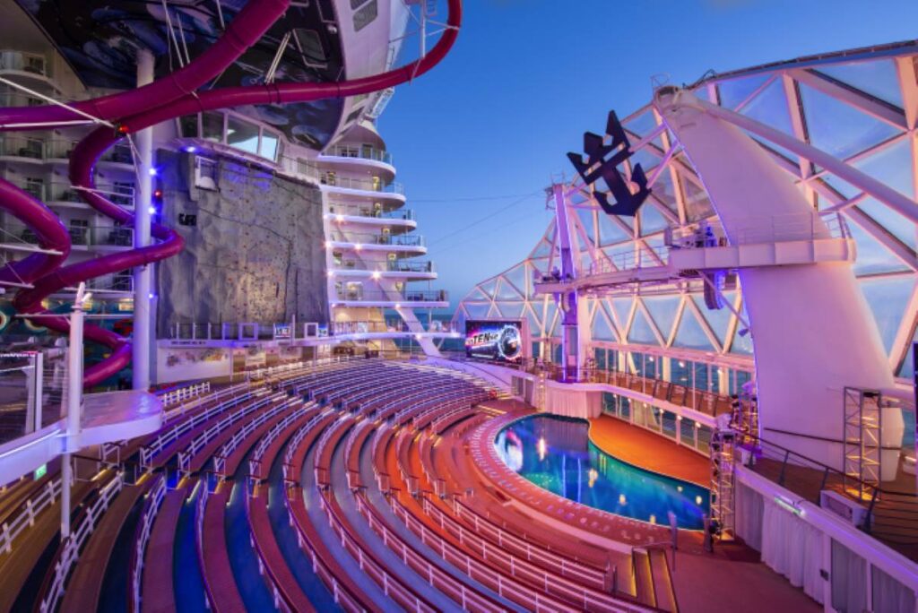 The AquaTheater on Wonder of the Seas captured at dusk, featuring an impressive outdoor amphitheater with tiered seating, a pool stage for aquatic shows, and a backdrop of colorful water slides and a rock climbing wall, all set against the ocean horizon.