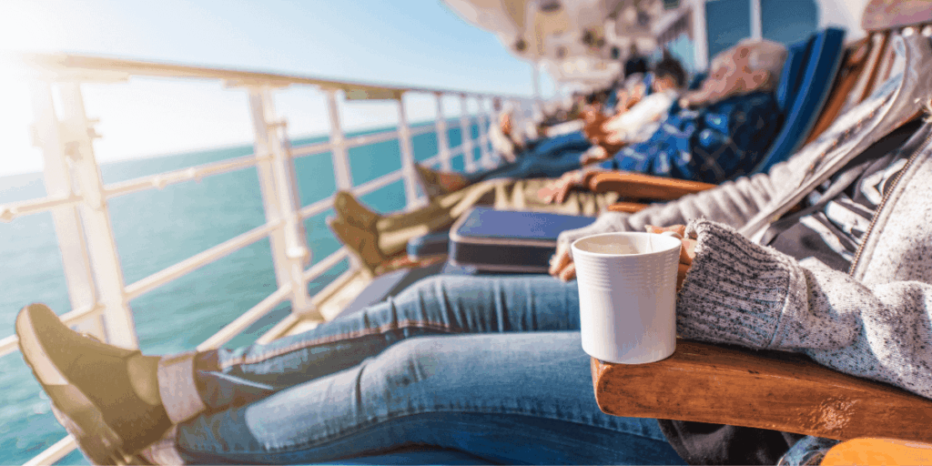 People relaxing in lounge chairs on a cruise ship deck, with one person holding a mug while ocean views stretch out beside the railing.