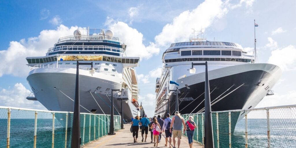 Cruise passengers walking down a pier toward two large cruise ships docked side by side under a bright, cloudy sky.