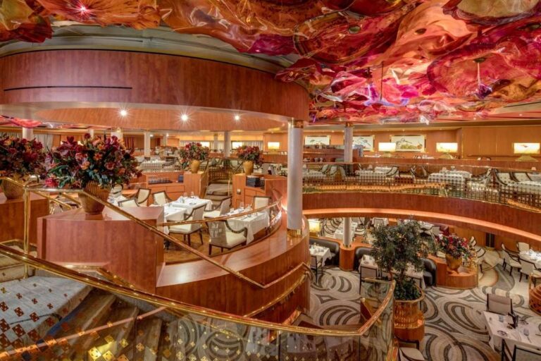 Two-level Holland America's Noordam main dining room with curved staircases, white tablecloths, warm wood finishes, and a colorful glass art ceiling.