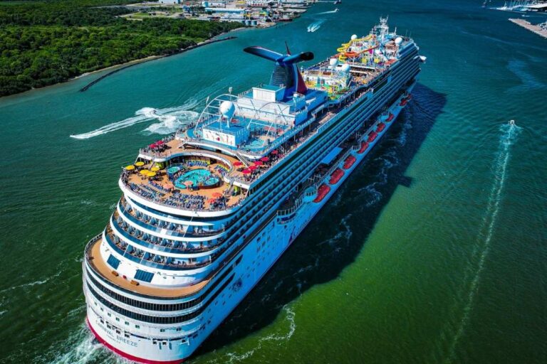 Aerial view of Carnival Breeze sailing through the Galveston channel, showing the ship’s upper decks, pool area, waterslides, and rows of lifeboats, with green shoreline and port facilities visible in the background.