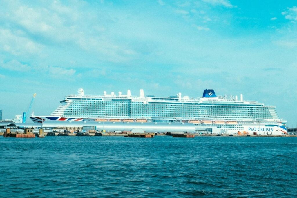 Large P&O Cruises ship docked at port beside calm blue water under a bright sky with scattered clouds.