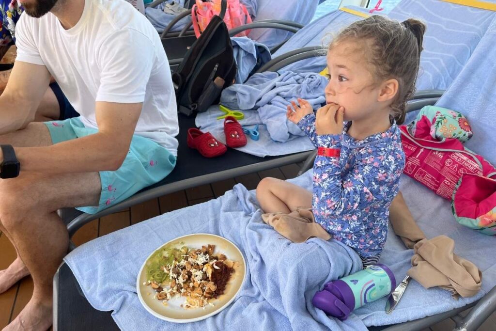 Child eating from a plate while sitting on a towel-covered pool lounger on a cruise ship deck.
