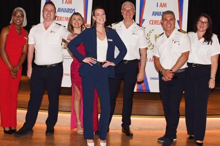 Sara Taskoski stands front and center in a navy suit during a Carnival onboard awards ceremony, posing with several uniformed crew members on stage.