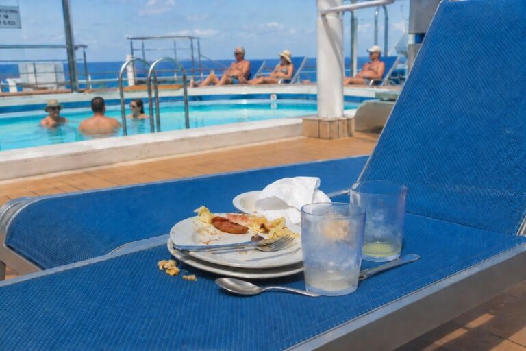 Dirty plates, cups, and utensils left on a blue cruise ship pool lounger while guests relax in the pool behind it.