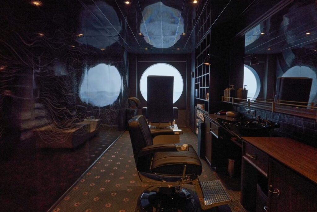 Dark, intimate interior of the Stubble & Groom barbershop, with black barber chairs, wood countertops, grooming stations, and round windows facing the ocean.