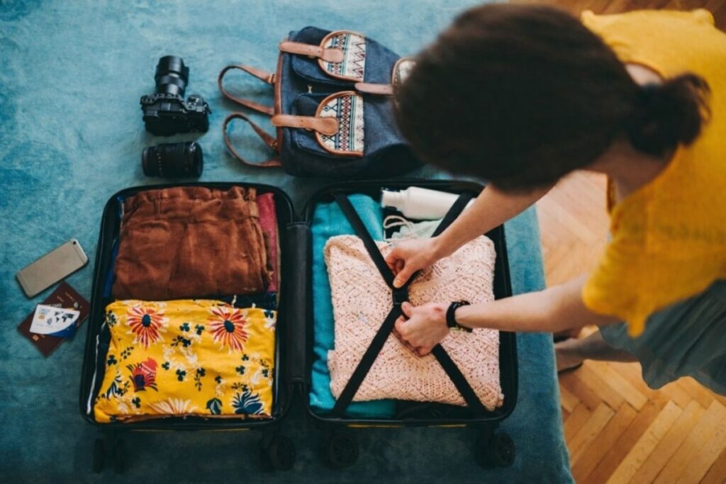 Person packing a suitcase with neatly folded clothes, toiletries, and travel essentials laid out beside an open carry-on bag.
