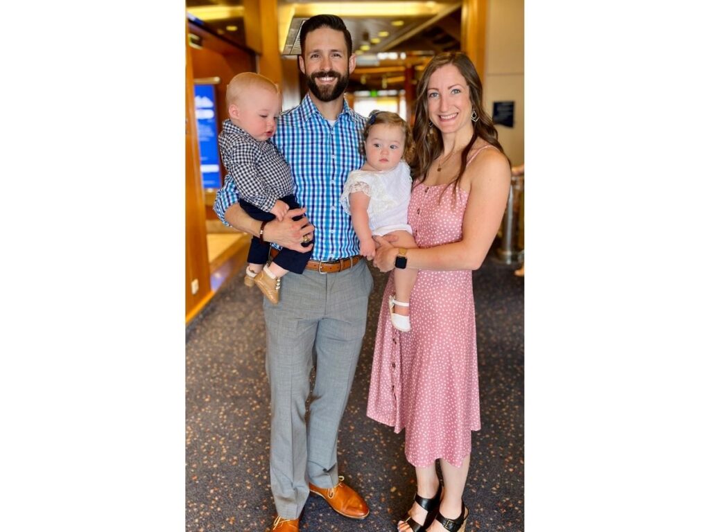 Family of four posing inside a cruise ship corridor, with a man holding a young boy and a woman holding a baby girl, all dressed in smart clothes for formal night.