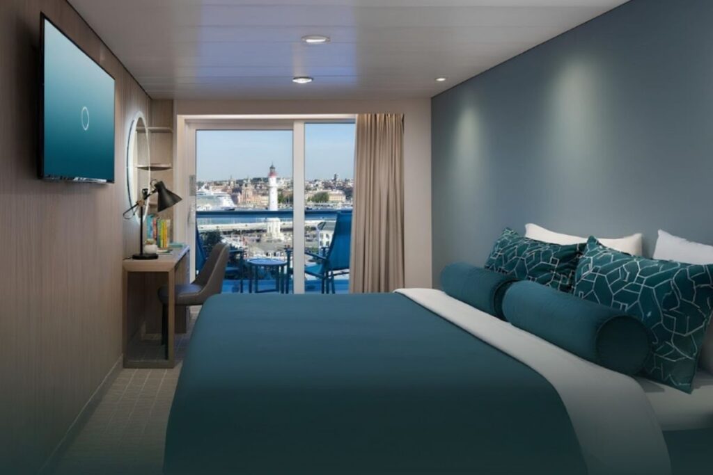 Modern cruise ship balcony cabin with teal bedding, a wall-mounted TV, desk and chair, and sliding glass doors opening to a private balcony with port views and a lighthouse in the distance.