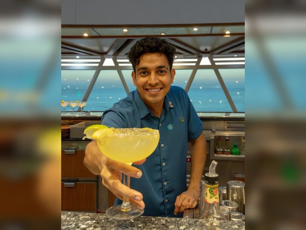 Cruise ship bartender smiles and holds out a margarita across the bar with ocean views in the background.