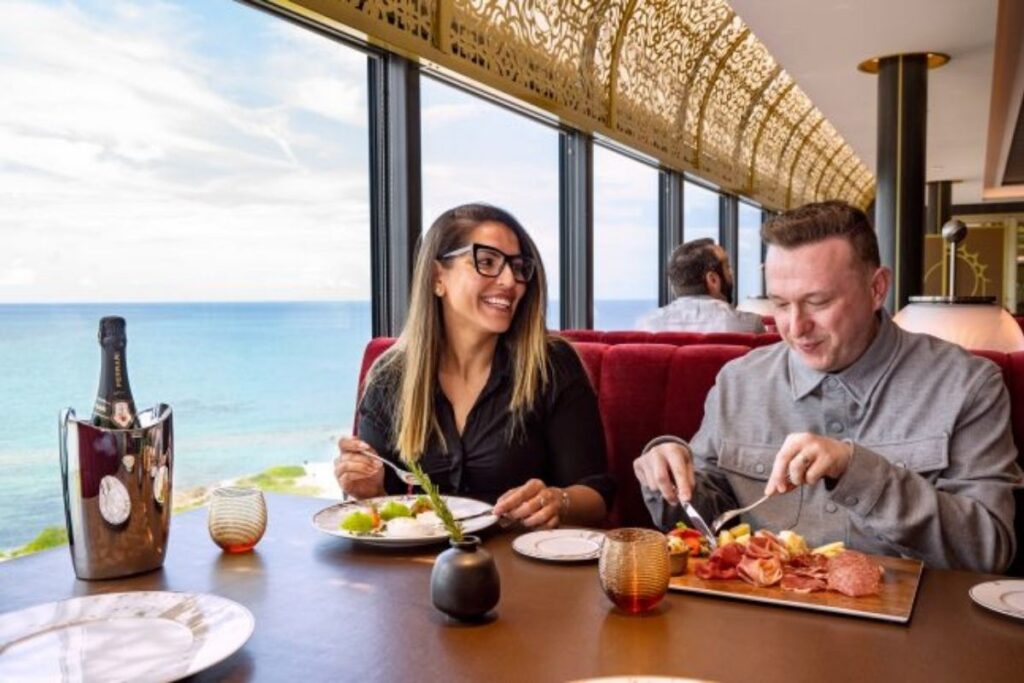 A couple sit at a Disney cruise ship restaurant table beside large windows overlooking the ocean, eating a plated meal with charcuterie, vegetables, and sparkling wine.