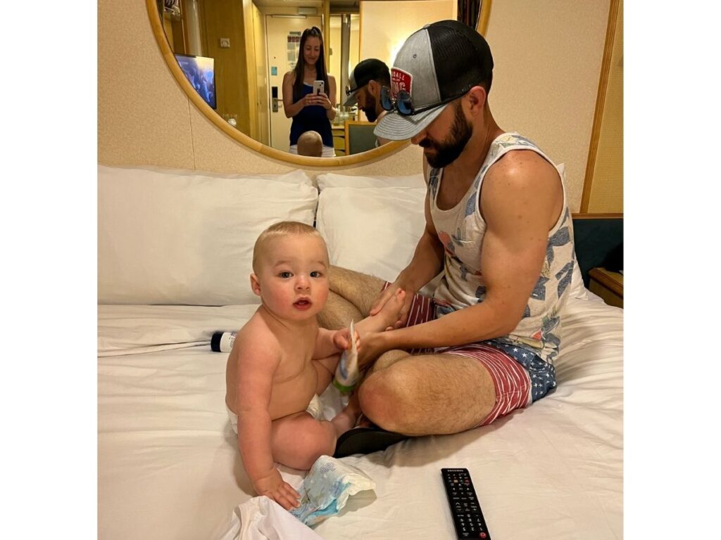 A father sits on a cruise ship cabin bed changing a baby’s diaper while the mother takes a photo in the mirror behind them.